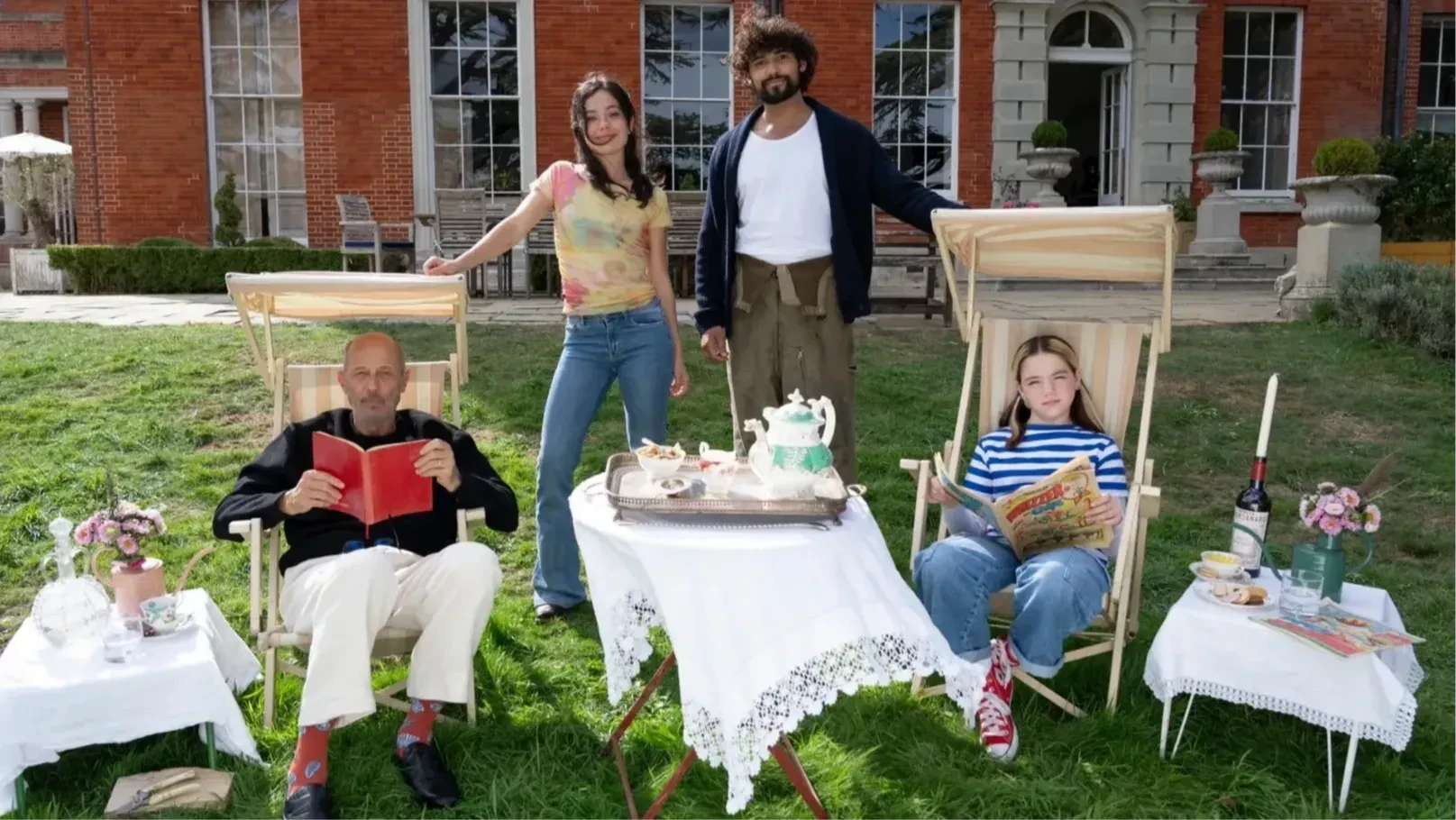 A group of people seated and standing on a lawn in front of a large house are looking directly into the camera.