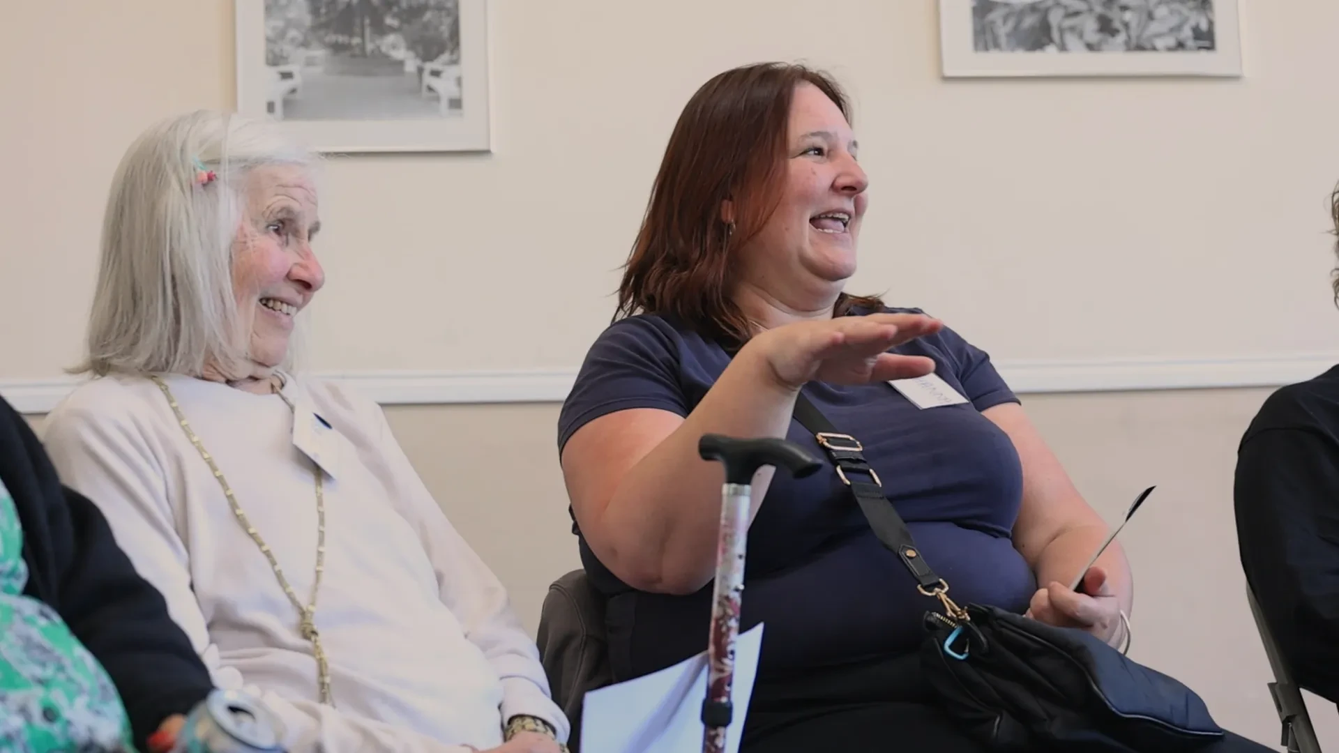 An older woman smiles as a younger woman sat next to her appears to be speaking.