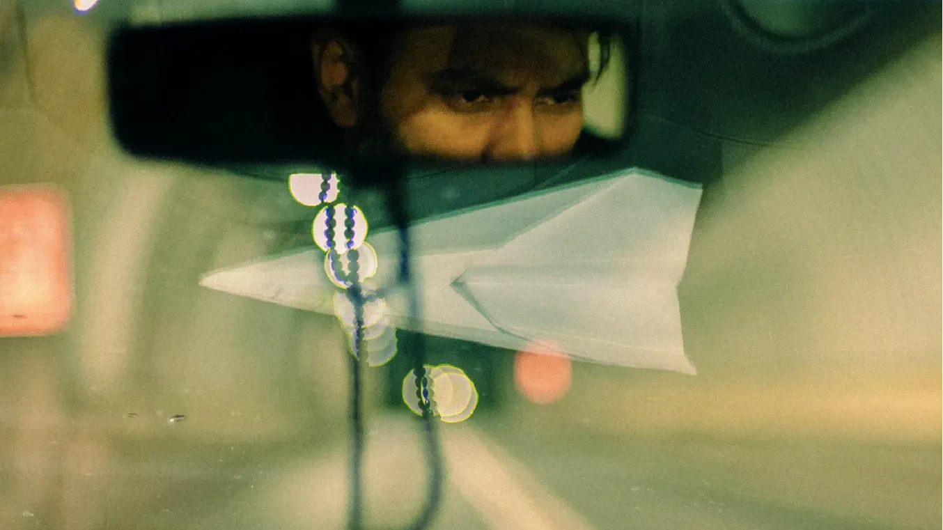 A ride-share driver looking in his rear view mirror, concentrating on the road ahead; a paper plane on the dashboard reflected in the window