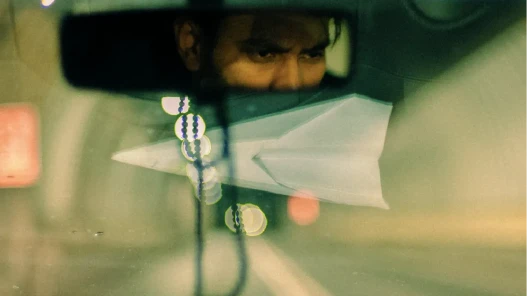 A ride-share driver looking in his rear view mirror, concentrating on the road ahead; a paper plane on the dashboard reflected in the window