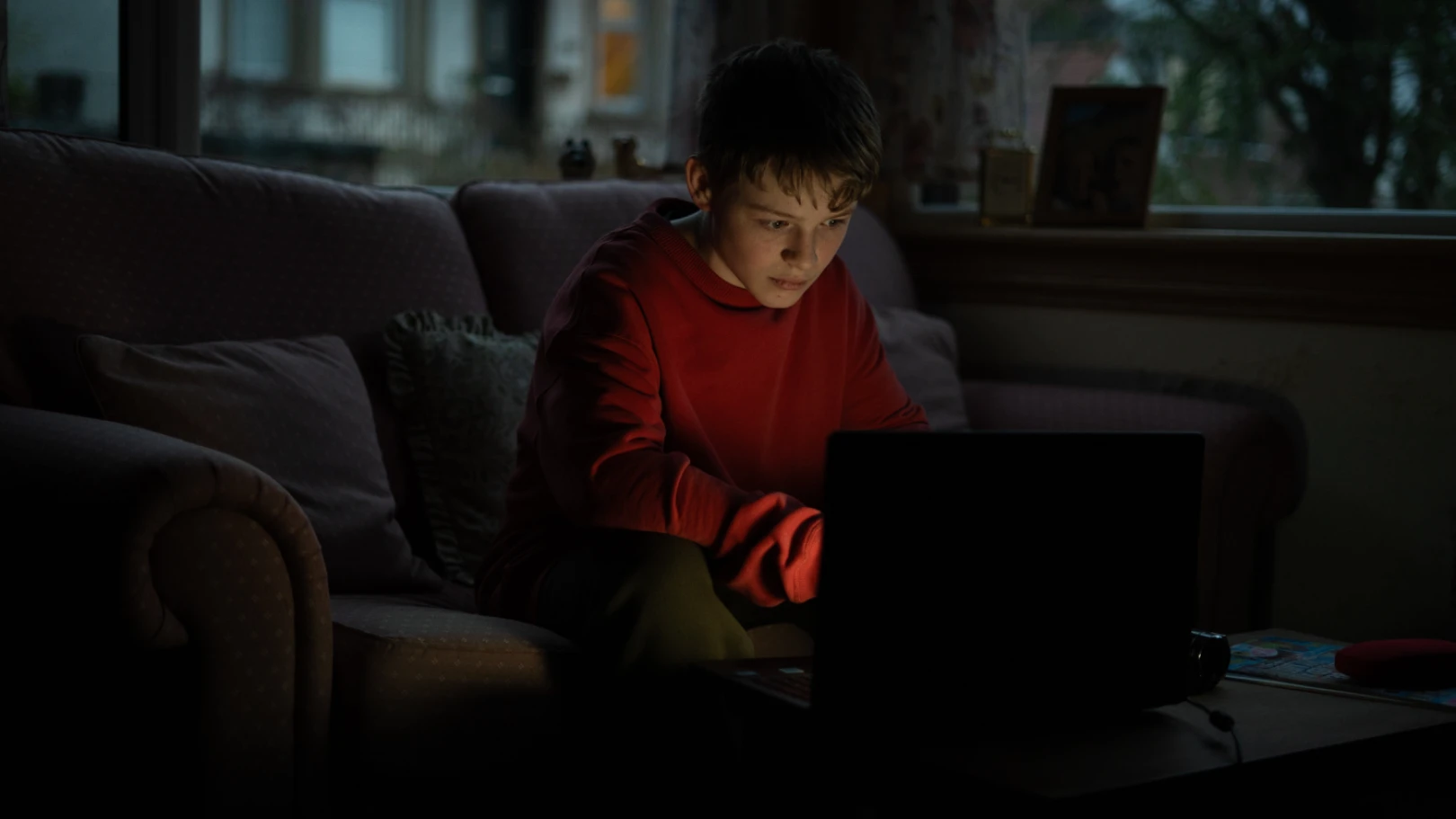A young teenage boy sits in front of a laptop at dusk in the living room with the screen lighting his face in the dark