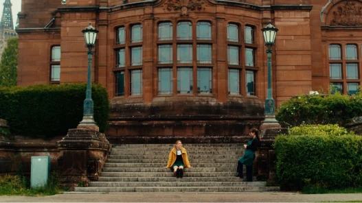 A young woman, wearing a bright yellow jacket, sits on the steps in front of the Kelvingrove Art Gallery and Museum in Glasgow.