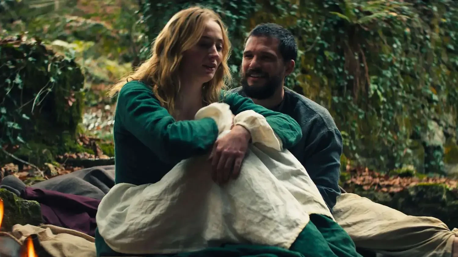 A woman in green sits in a forest clutching her knees while a man looks on smiling