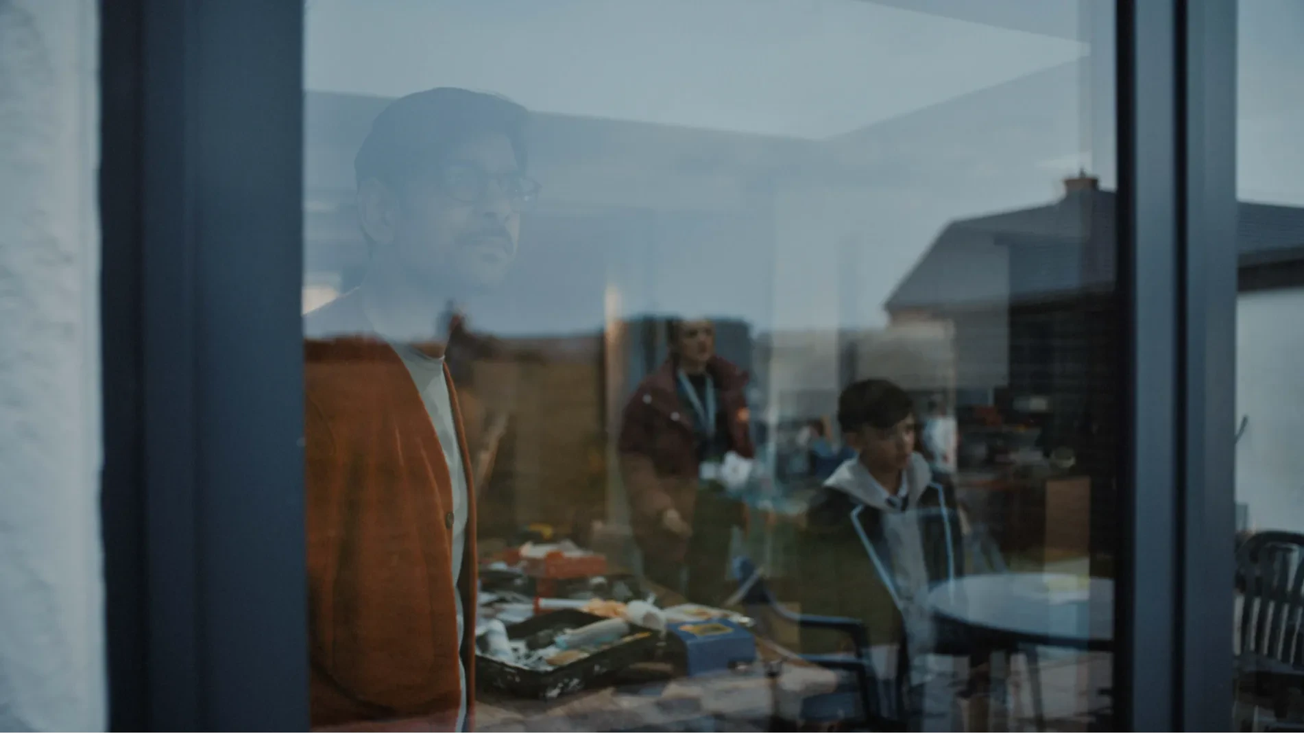 A family look out from their kitchen into their garden. We see them through the glass from the outside.