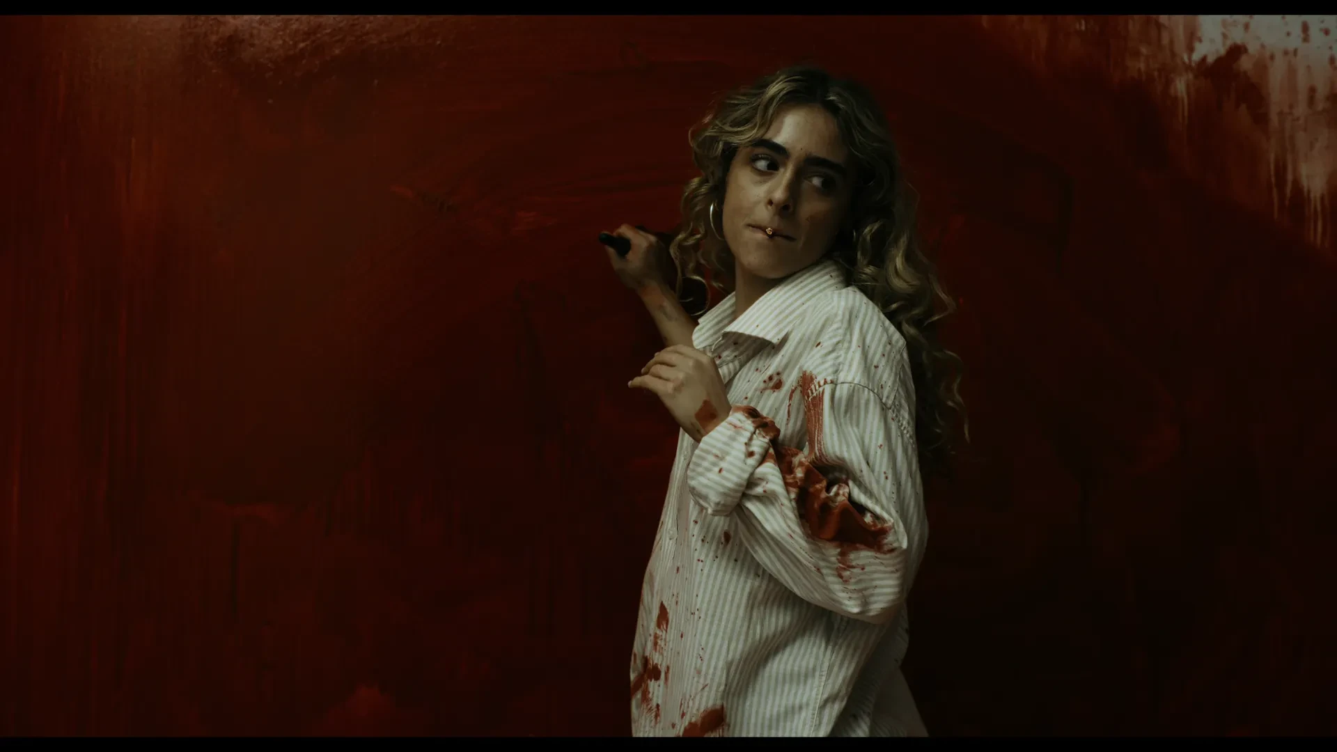 A woman with a cigarette in her mouth stands in front of a bright red wall