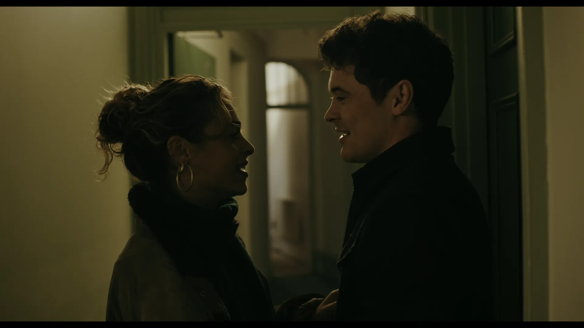 A young man and young woman stand close together in a corridor, staring into each other's eyes
