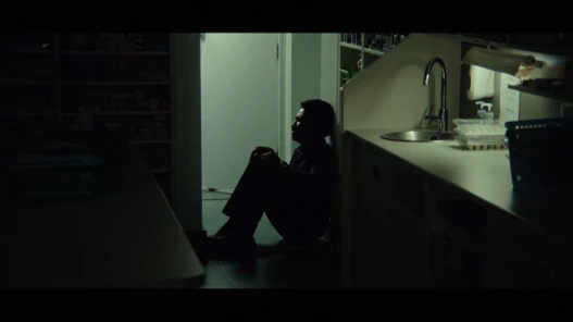 A young, black, male pharmacist sits on the floor of a dark dispensary holding his knees up to his chest