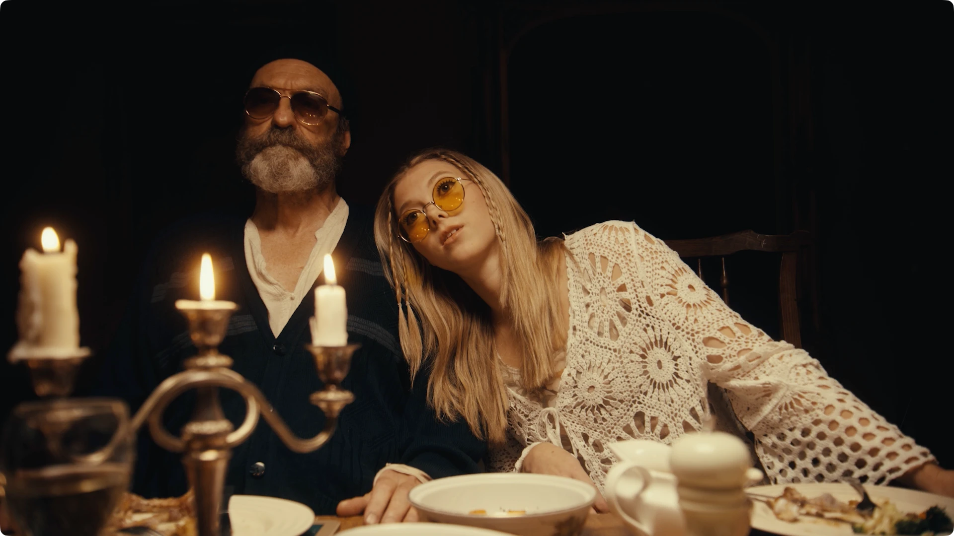 A male and female couple at dinner party behind a candelabra, both wearing sunglasses. The woman in white leans on the man in a suit.