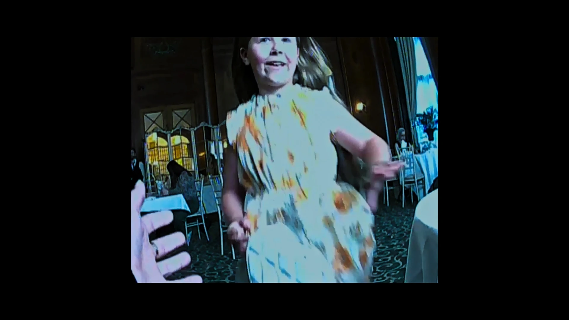 A recording of a smiling little girl in a white dress with yellow patterns running towards a person holding the camera