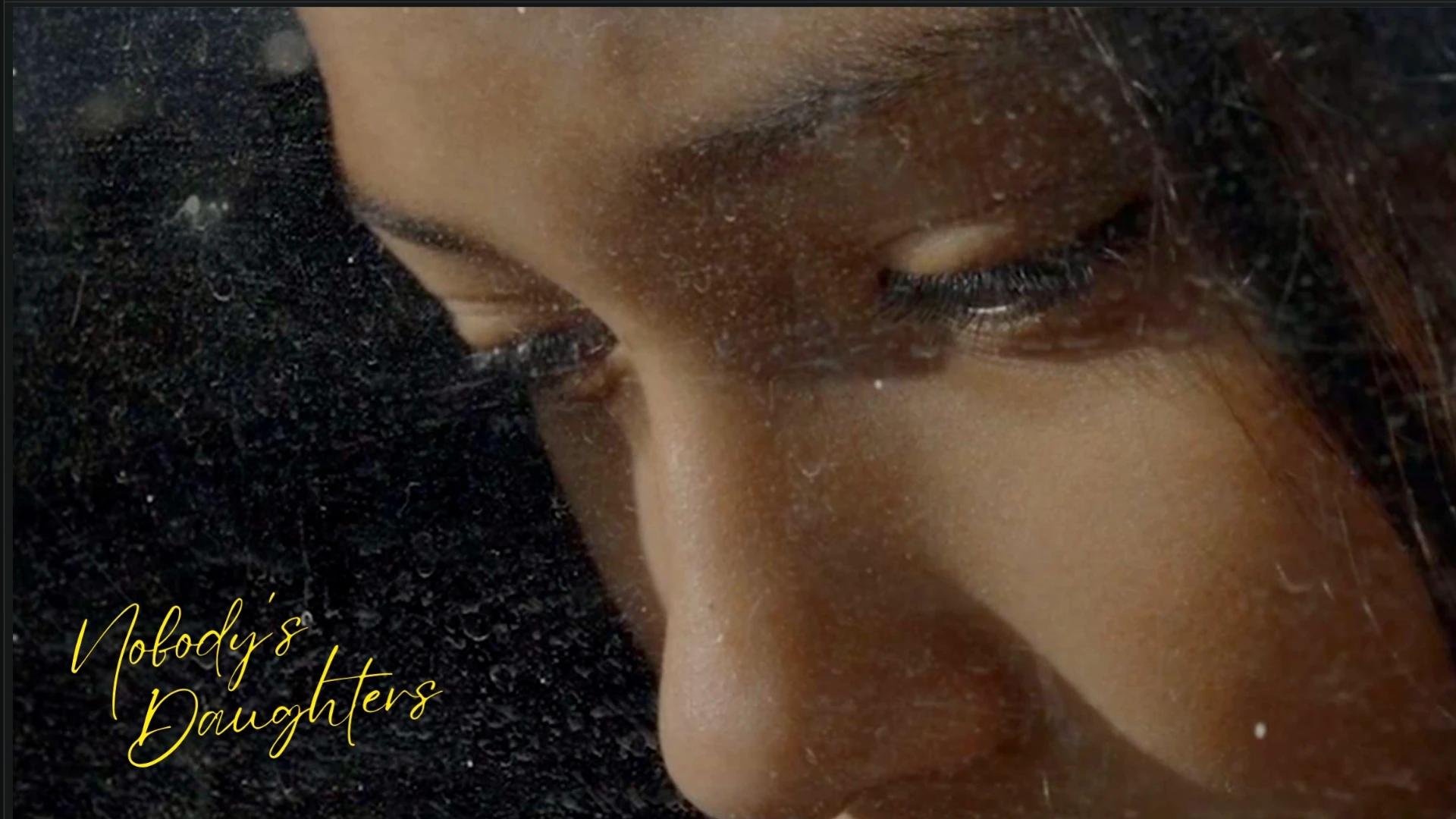 A close up of a woman looking down with a black background and "Nobody's Daughter" written in yellow