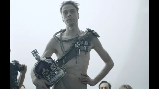 A man without a top on wears a crossbody sash with medals and holds an elaborate crown