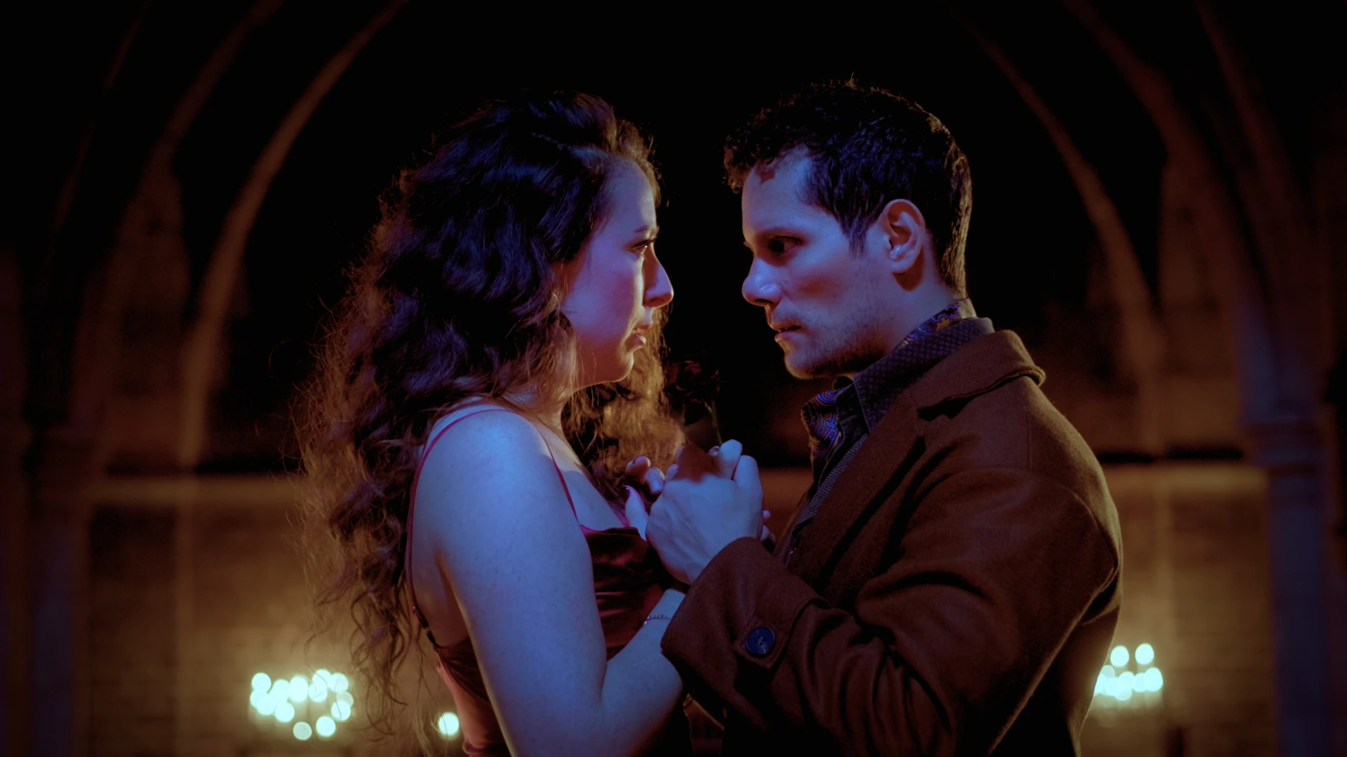 A man and a woman standing in front of each other under the arch of the candle lit cathedral. Their expressions depict passion and tension.