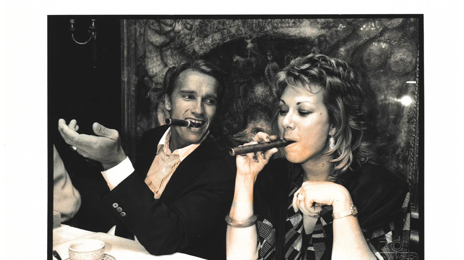 A black and white archive image of Arnold Schwarzenegger smoking a cigar with a woman