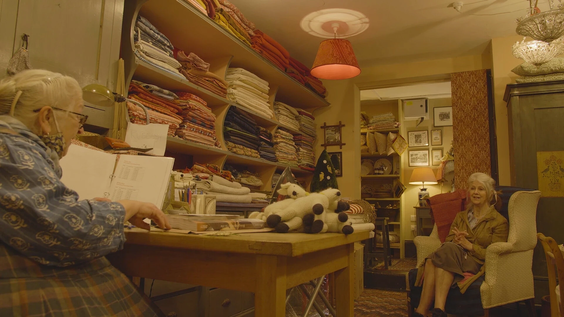A lady with white hair sat in a chair talking with a customer surrounded by fabrics