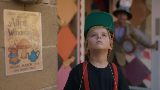 A young boy looks upwards. Behind him is a poster advertising 'Alice in Wonderland'