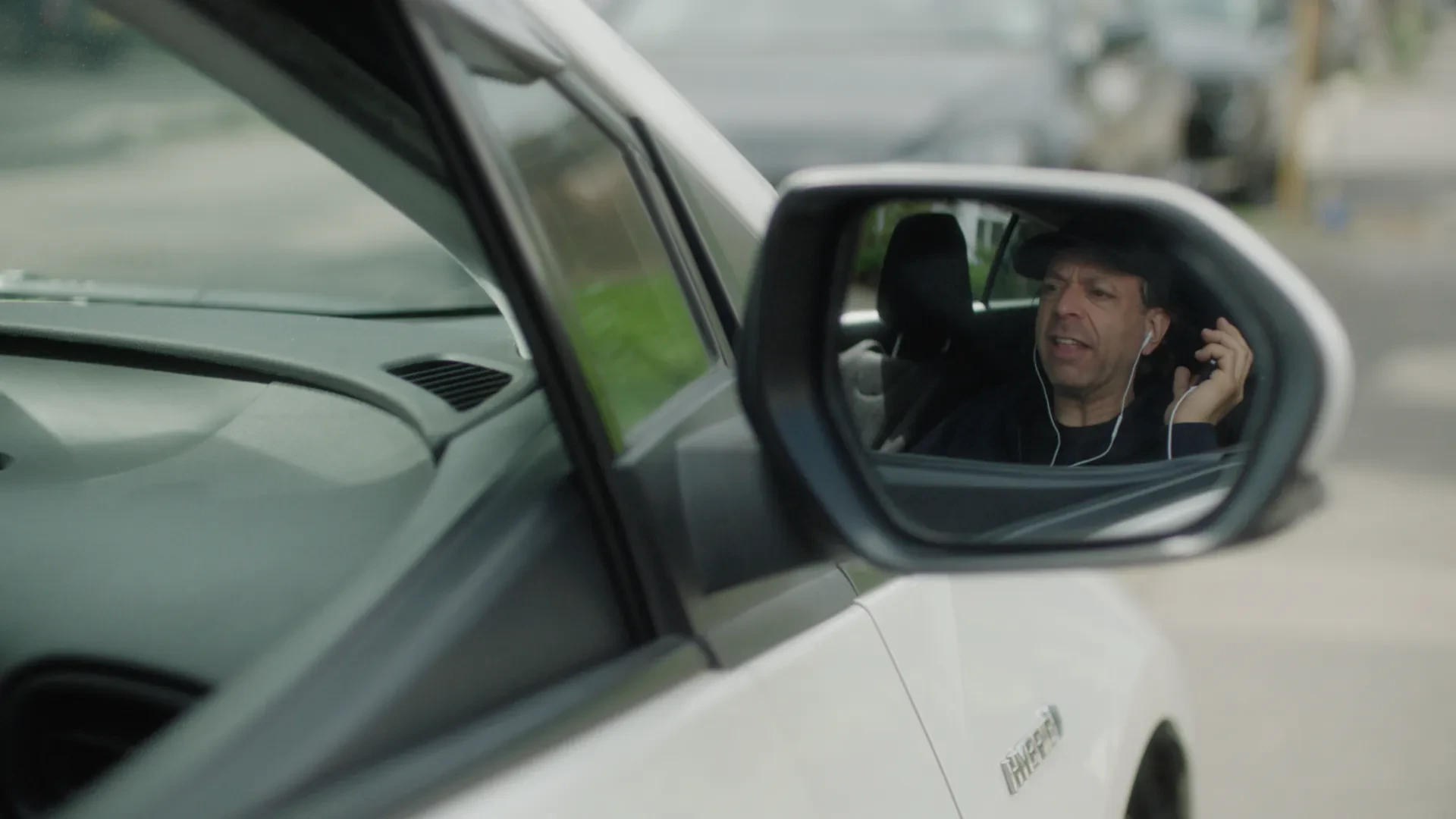 Through the reflection of a car's side mirror, man in a baseball hat is having a phone call