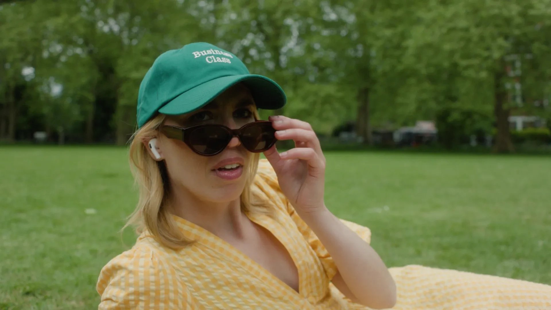 A woman in a green baseball hat and yellow dress pulling down her glasses in surprise. 