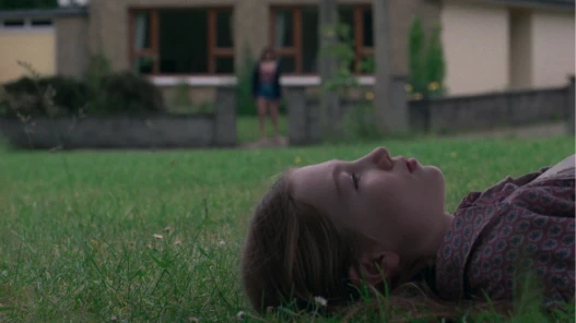 A young girl is lying on the grass staring up at the sky in front of her house, in the background her mother stands at the front gate of their house looking towards her.