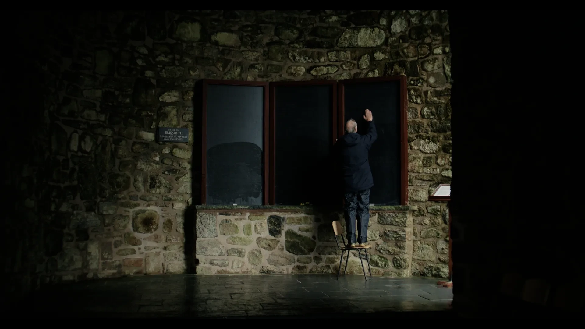 A man stood on a chair wipes water vapour with his hand off the glass cover of a memorial board in a stone brick chapel.