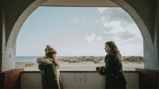 A girl and a woman talk with a view of a beach and the sea behind them.