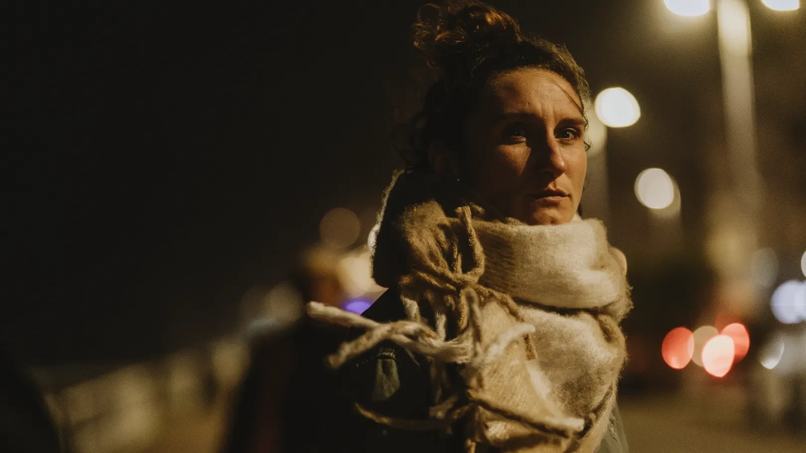 Woman wearing a large scarf outdoors at night, with blurred lights in the background.