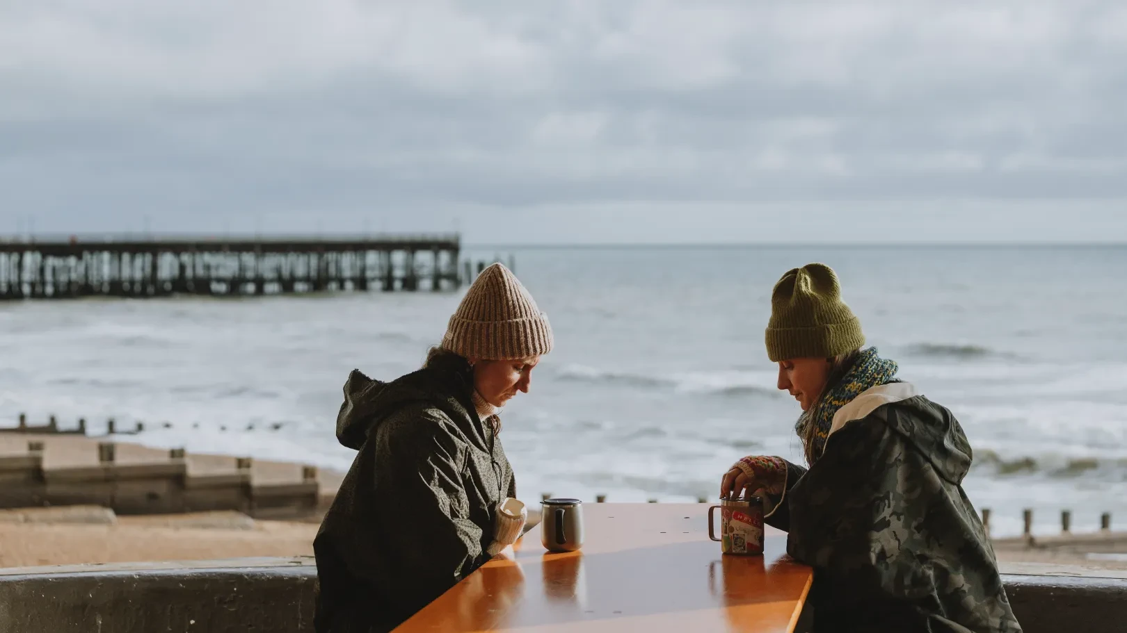 Two women in winter hats sitting with hot drinks at a table overlooking the sea and a pier.