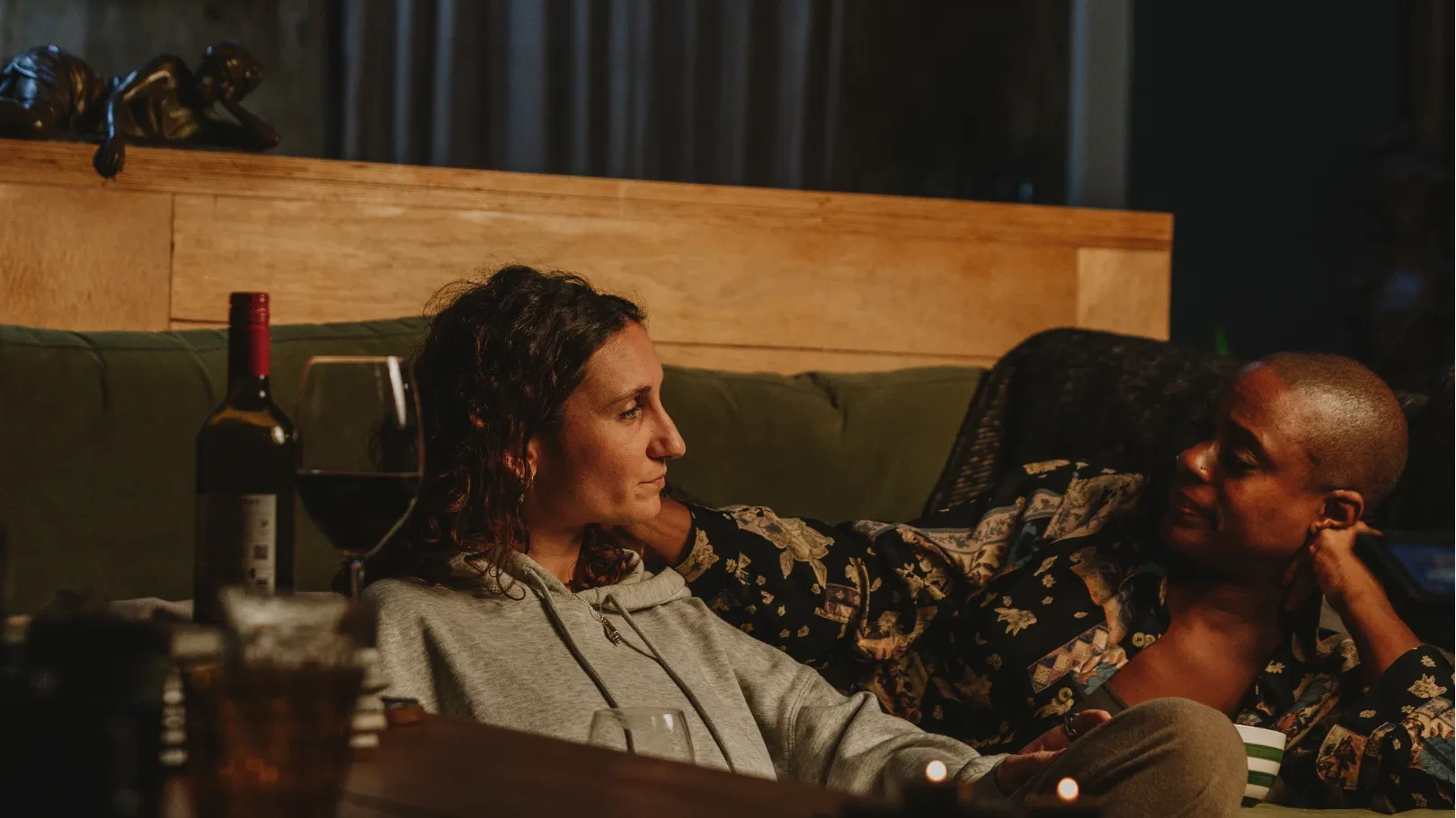 Two women relaxing together on a sofa, talking over drinks in a warmly lit room.