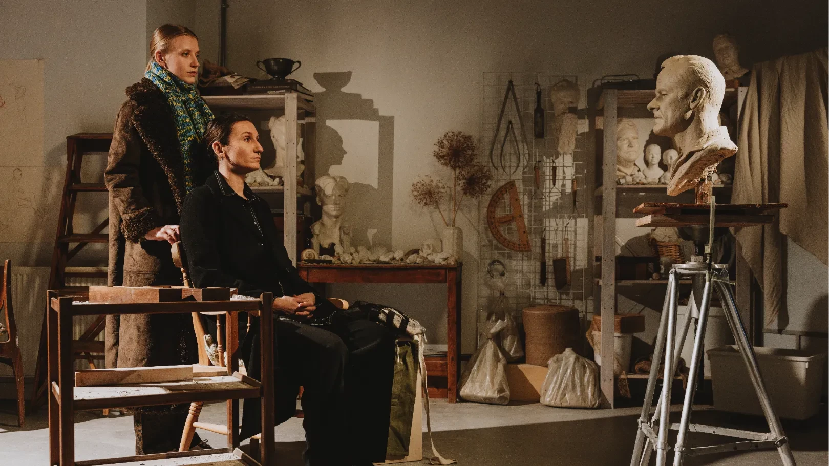 Two women looking at a sculpted bust of a man in an artist’s studio filled with sculptures and tools.