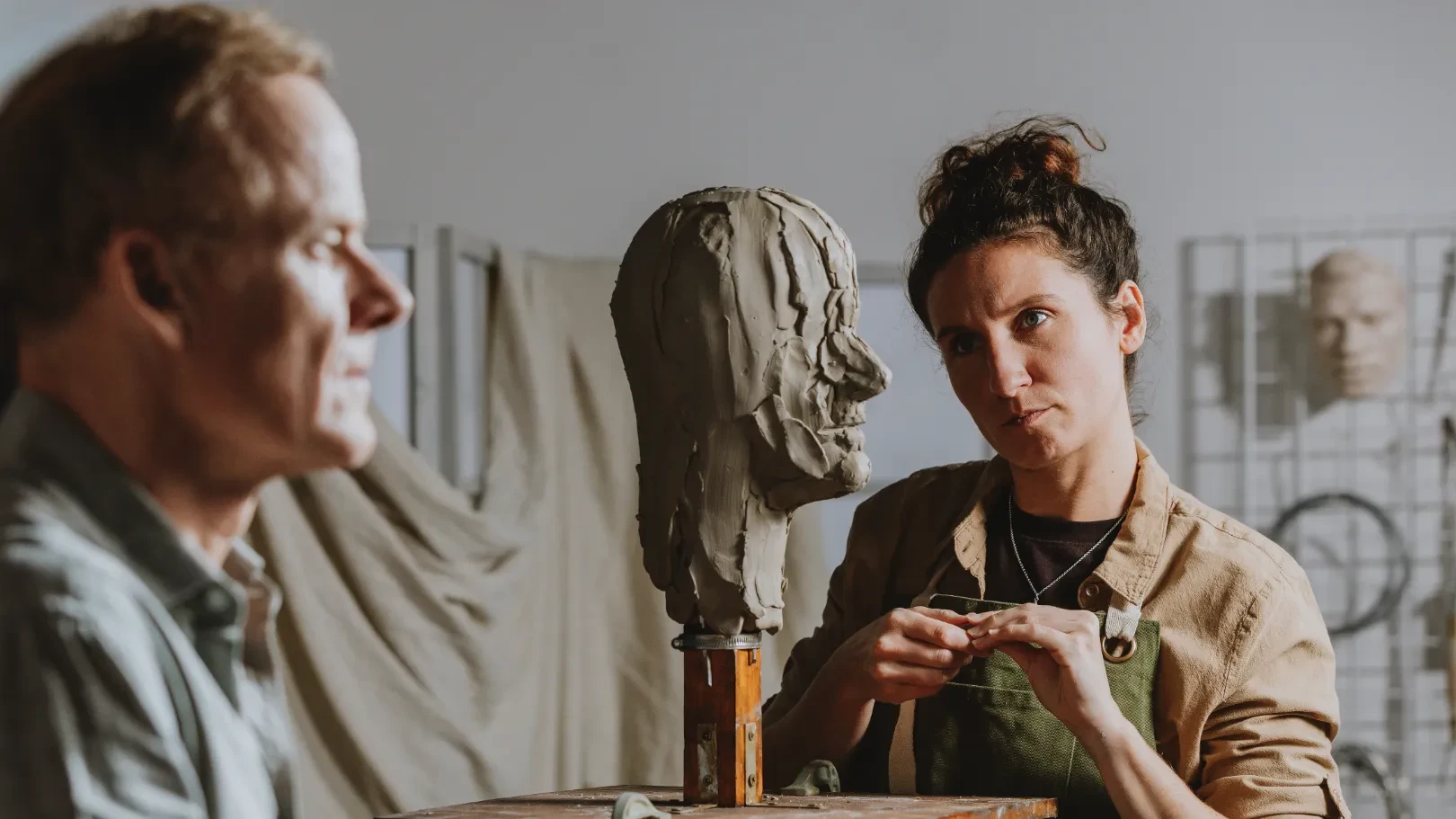 Woman sculpting a clay bust in a studio, with a man seated out of focus in the foreground.