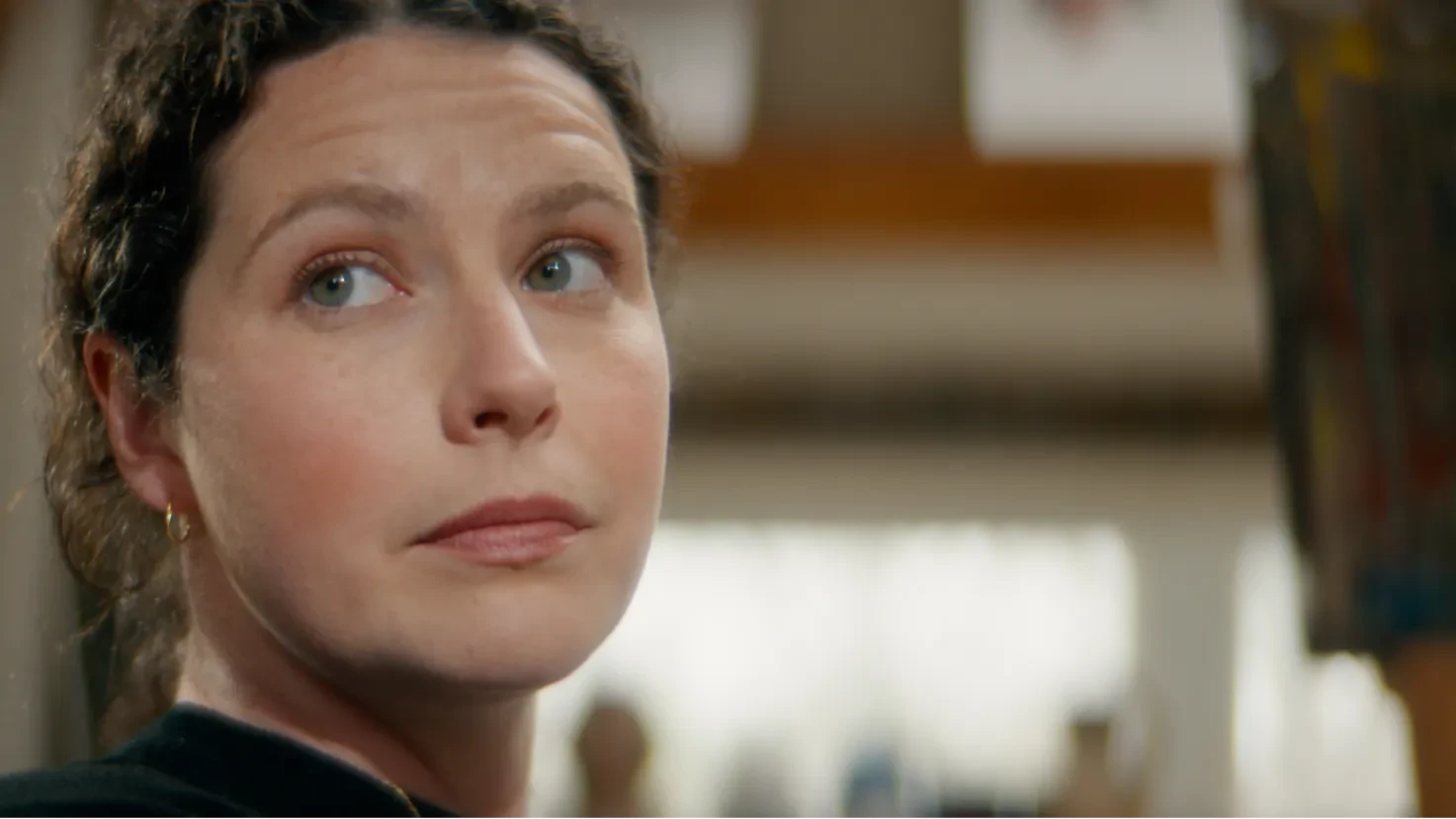 A woman with dark hair looks thoughtfully to the side in a warmly lit art studio.