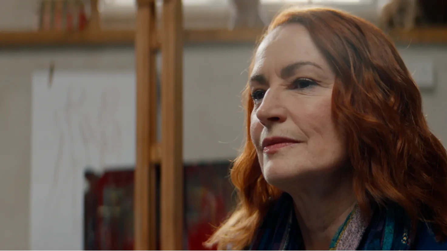 A woman with auburn hair looks thoughtfully to the side in a warmly lit art studio.