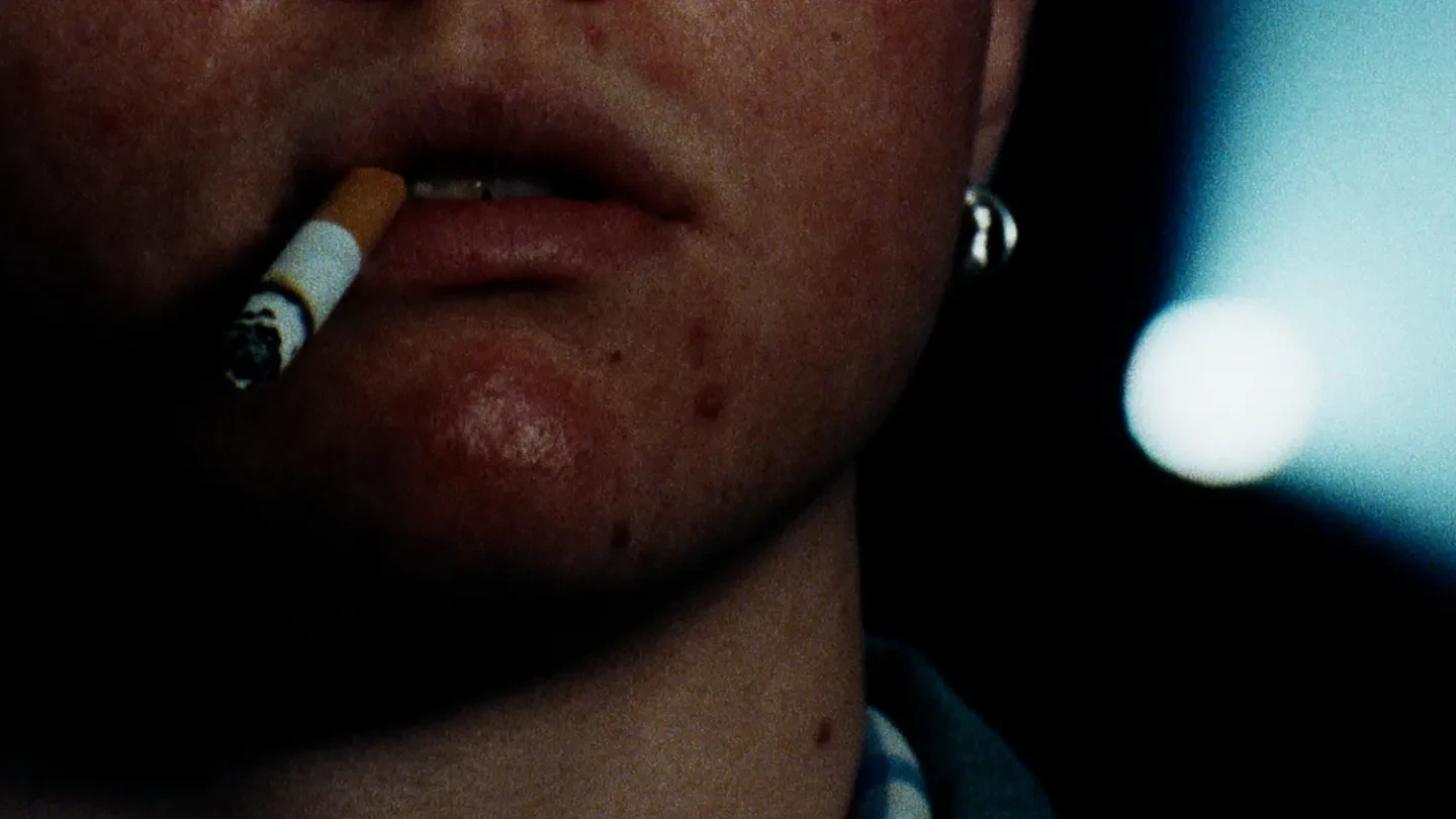 The lower-half of a person's face, with a cigarette in their mouth. A film projector in the dark background.