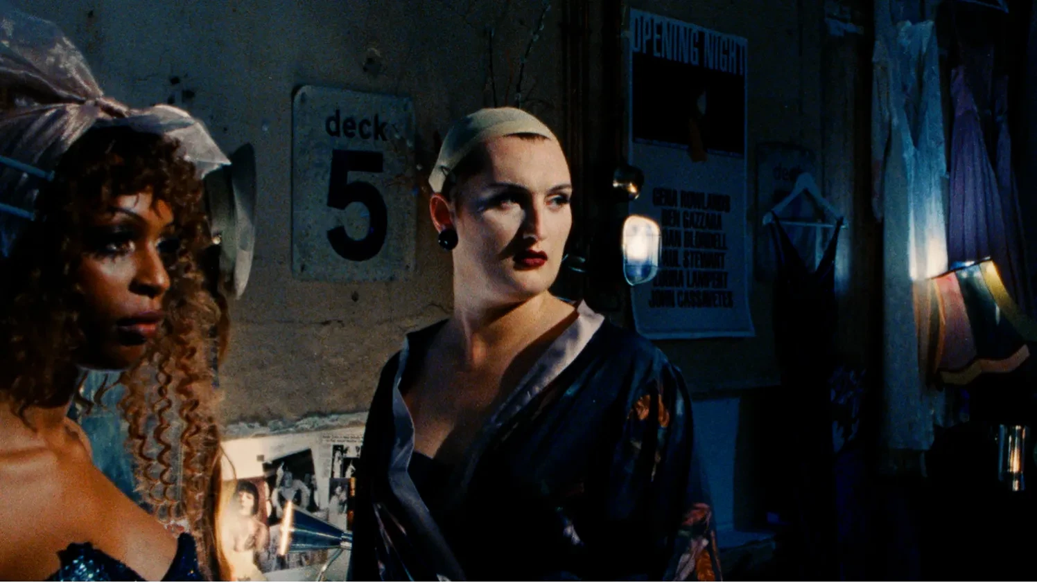 Two drag queens stand in a darkened dressing room