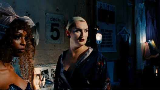 Two drag queens stand in a darkened dressing room