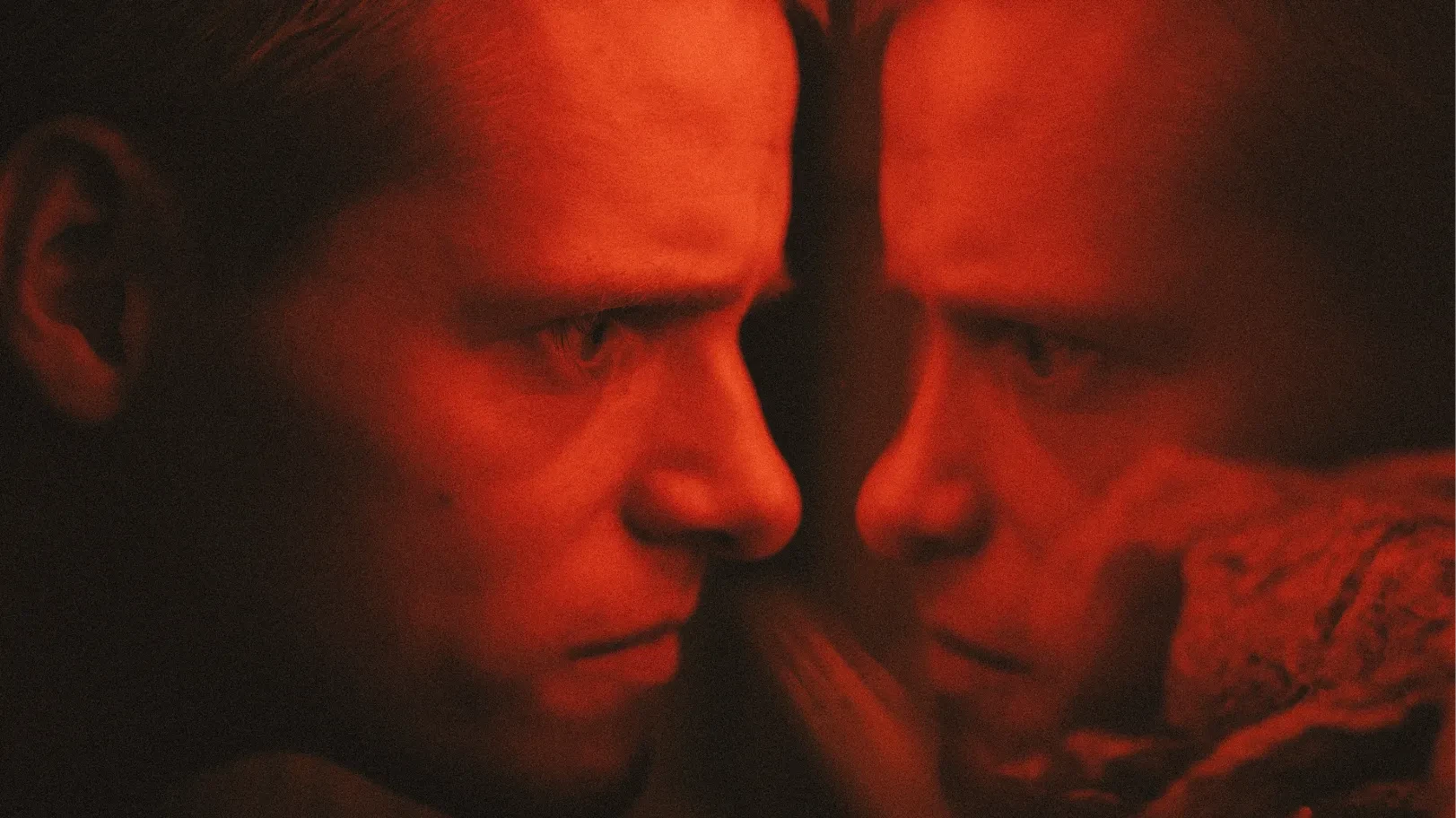 A young man bathed in red light from a reptile enclosure staring at his own reflection in the glass.