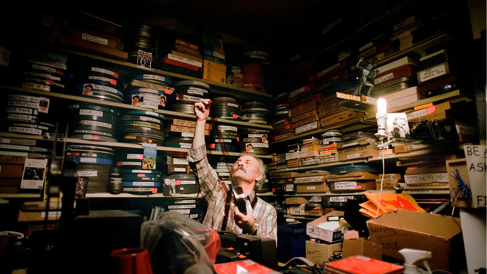 Umit Mesut holding a strip of film up to a light, surrounded by shelves packed with film canisters and movie memorabilia.