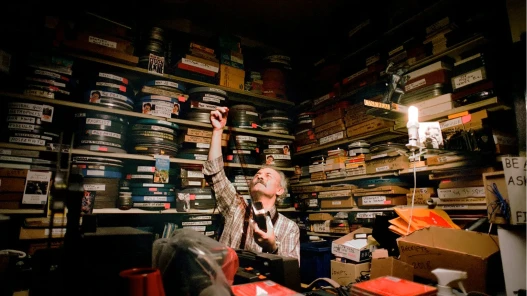 Umit Mesut holding a strip of film up to a light, surrounded by shelves packed with film canisters and movie memorabilia.