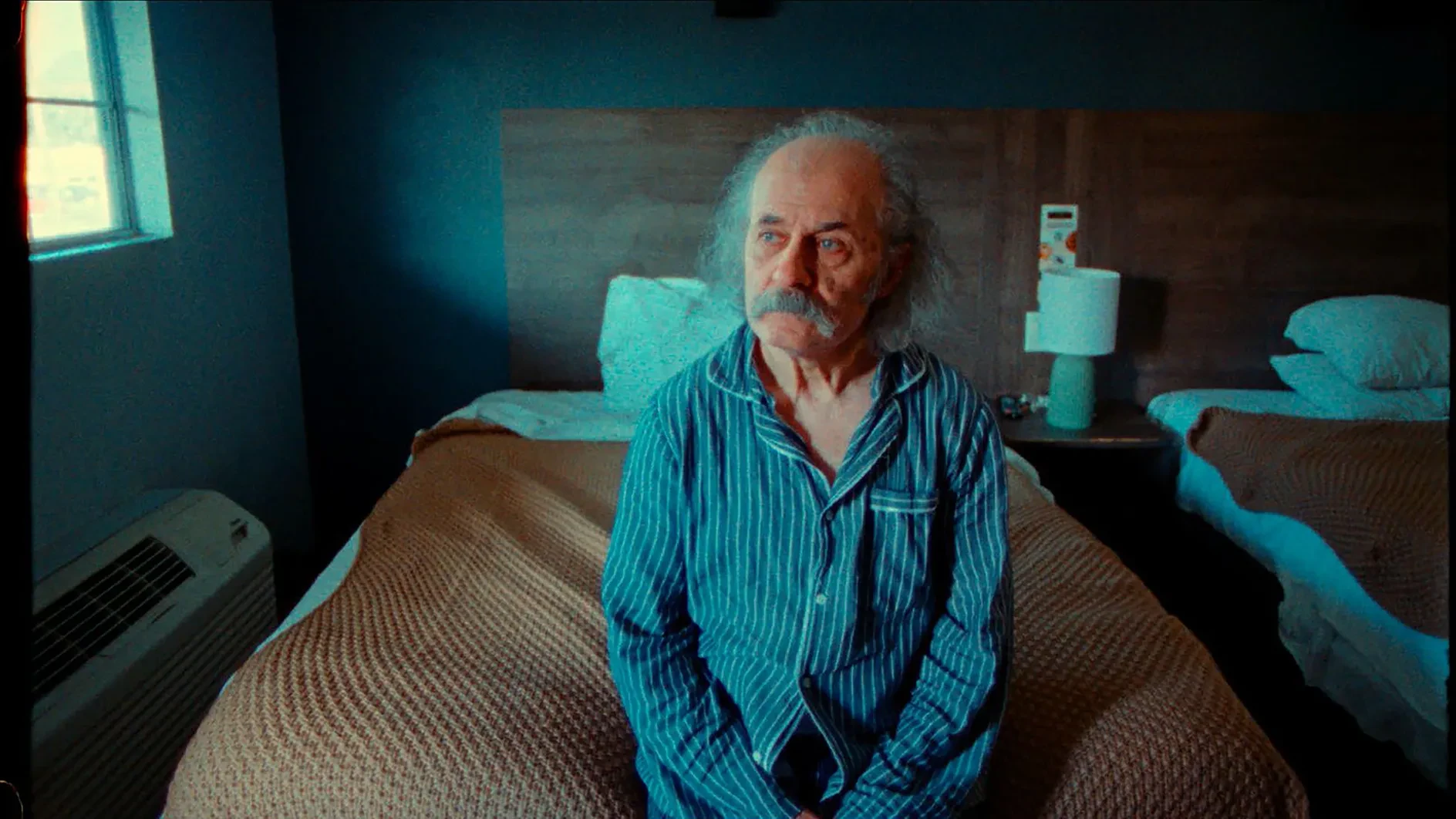 Umit Mesut in blue pinstriped pajamas sitting on the edge of a bed in a motel room.