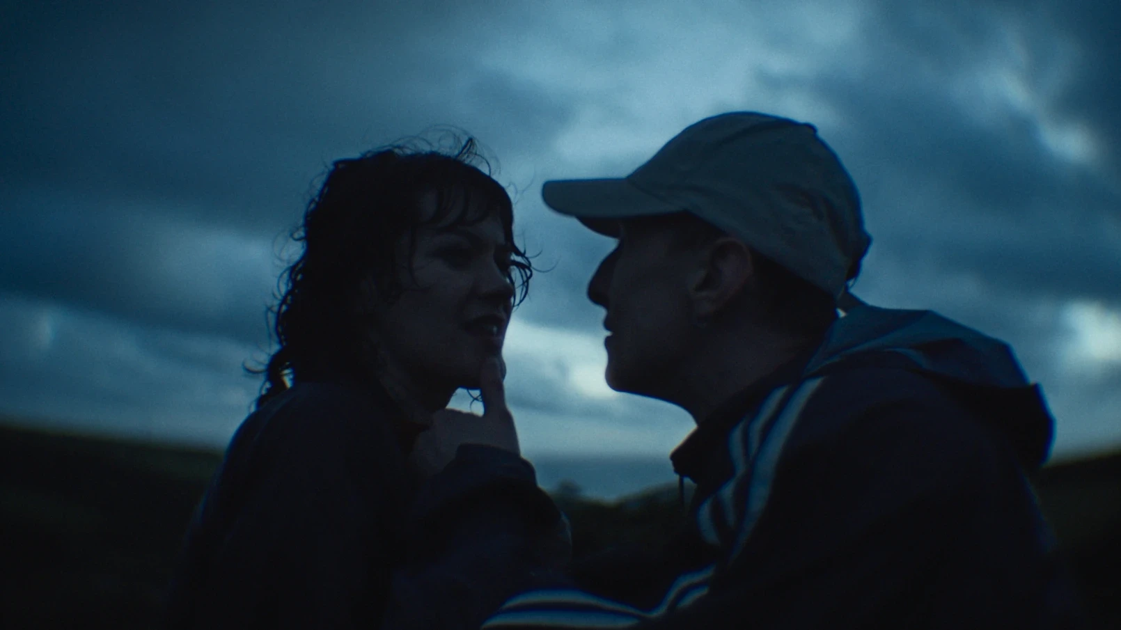 A man touches a woman's chin with is thumb as they stand against a cloudy sky.