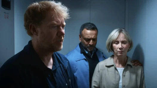 A man in a caretaker's uniform and two other adults, all looking sad, are standing in a lift.