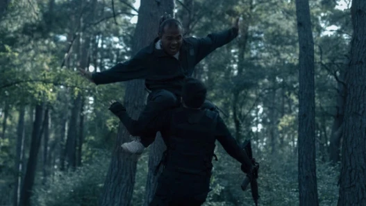 A man in a forest jumps screaming at another man in black holding a gun
