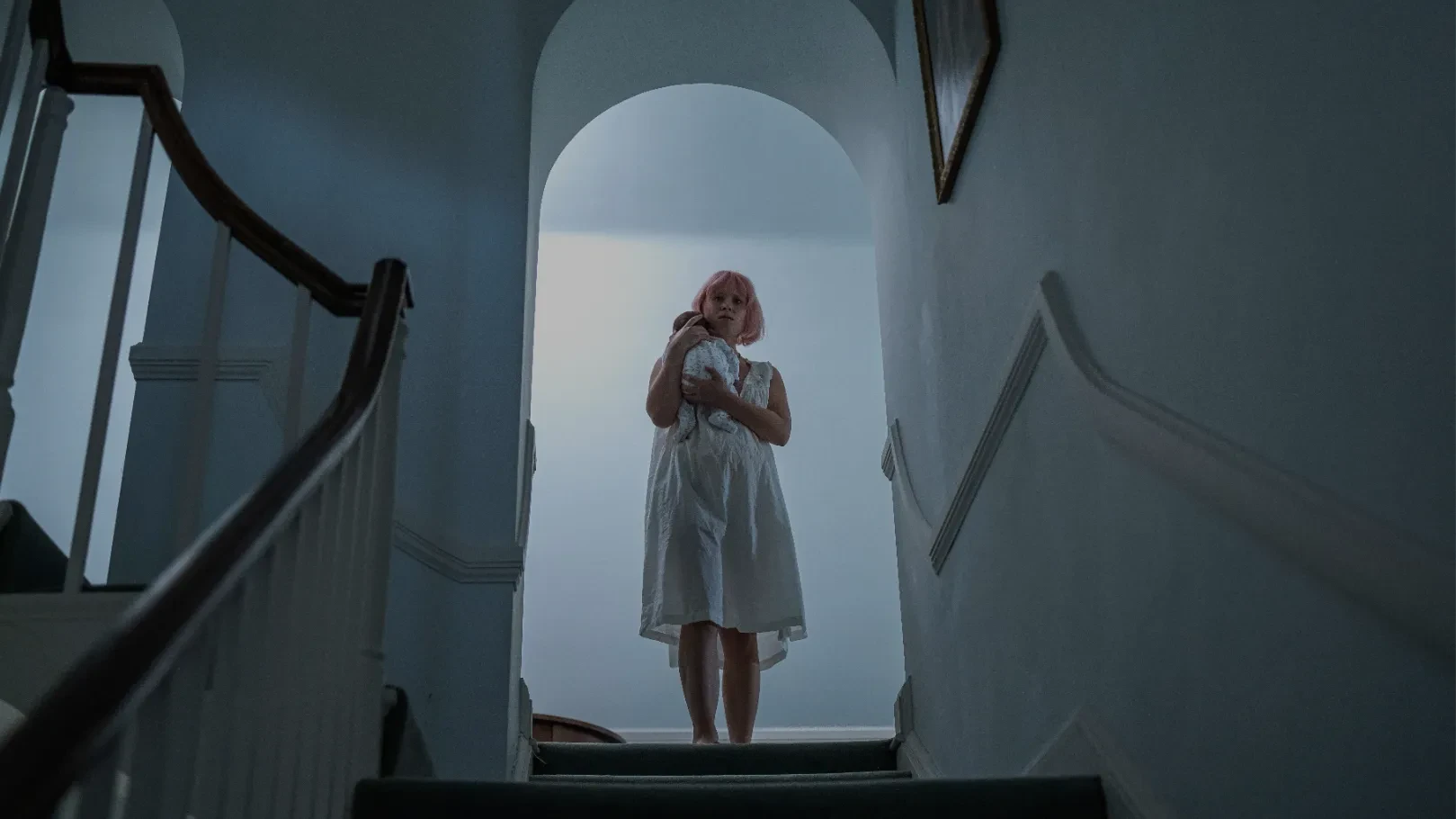 A blonde woman in a white dress holds a baby at the top of a staircase