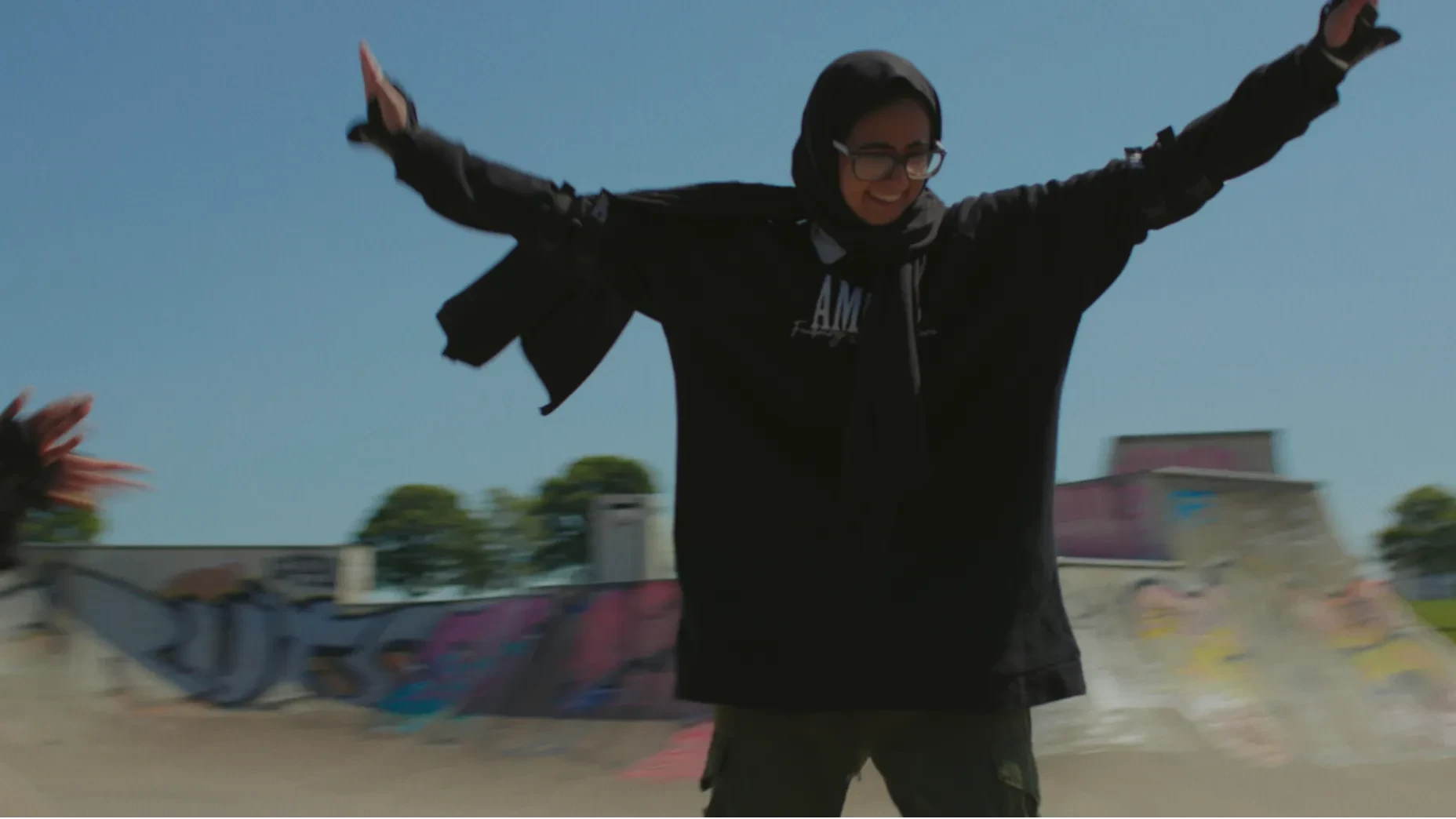 A young girl in a hijab, with arms wide open and a smile on her face, in a skatepark in daylight