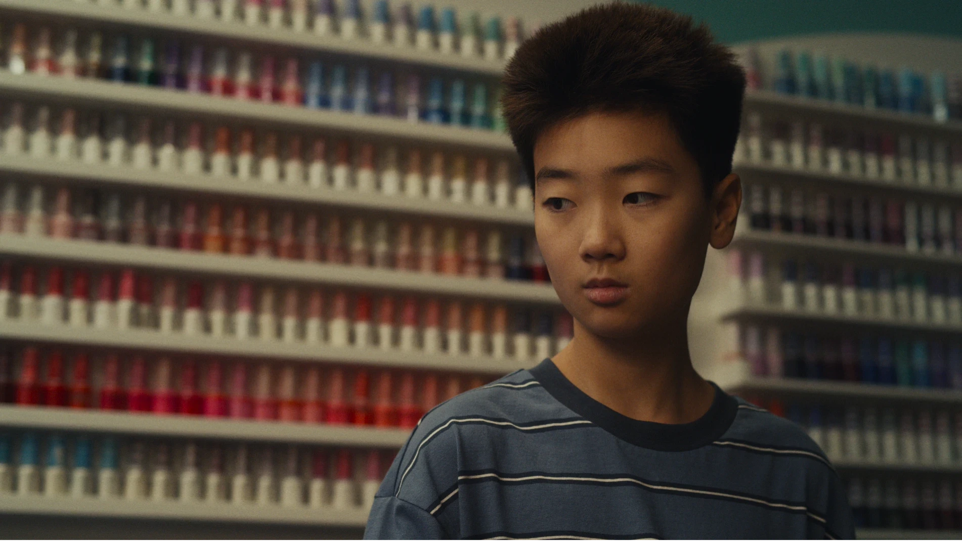 A young Asian boy wears a striped blue top and stands in front of rows of nail varnish bottles.