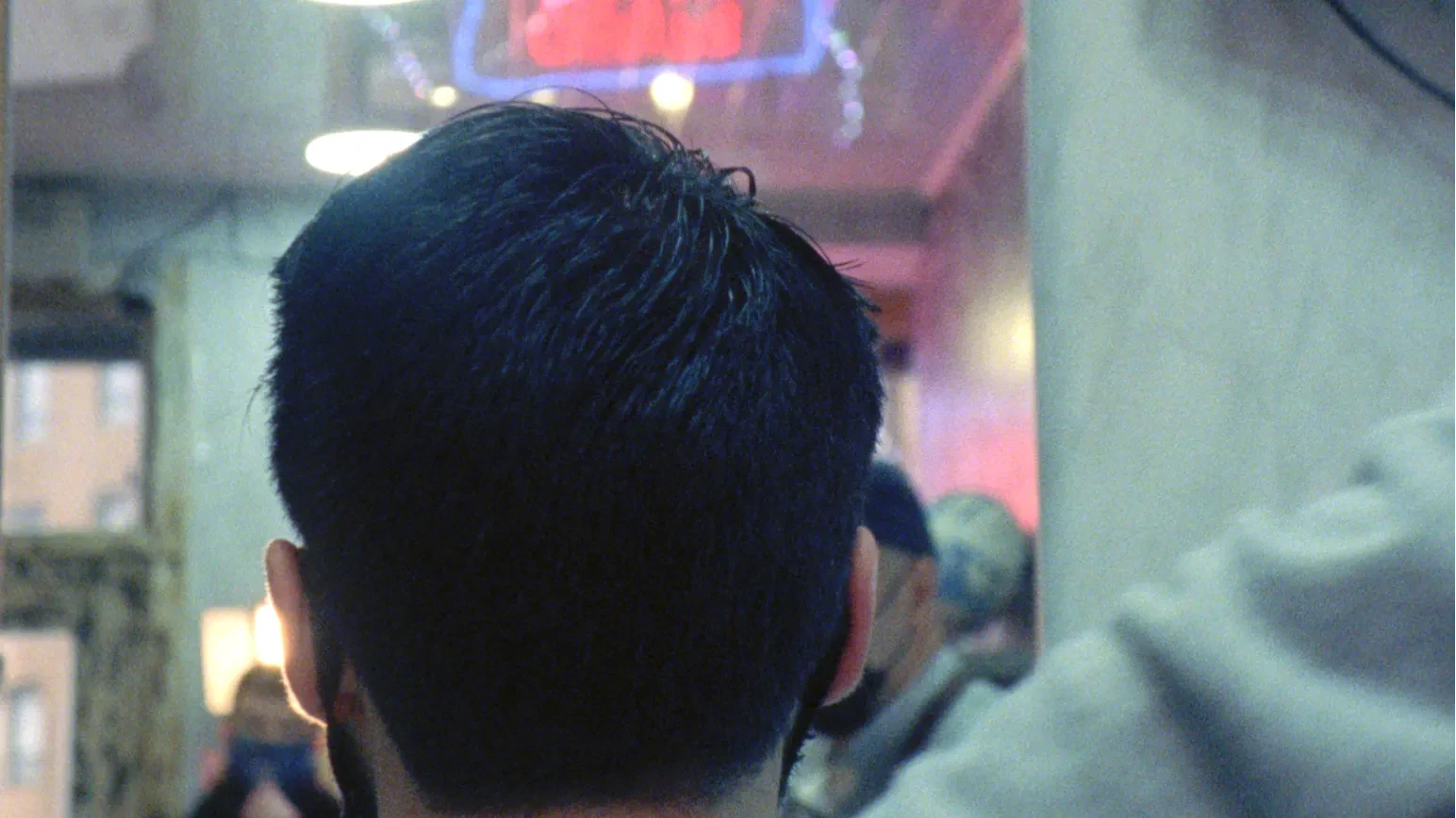A grainy, blue-green tinted film still showing the back of a person’s head in the foreground, facing into a softly lit, blurred interior.