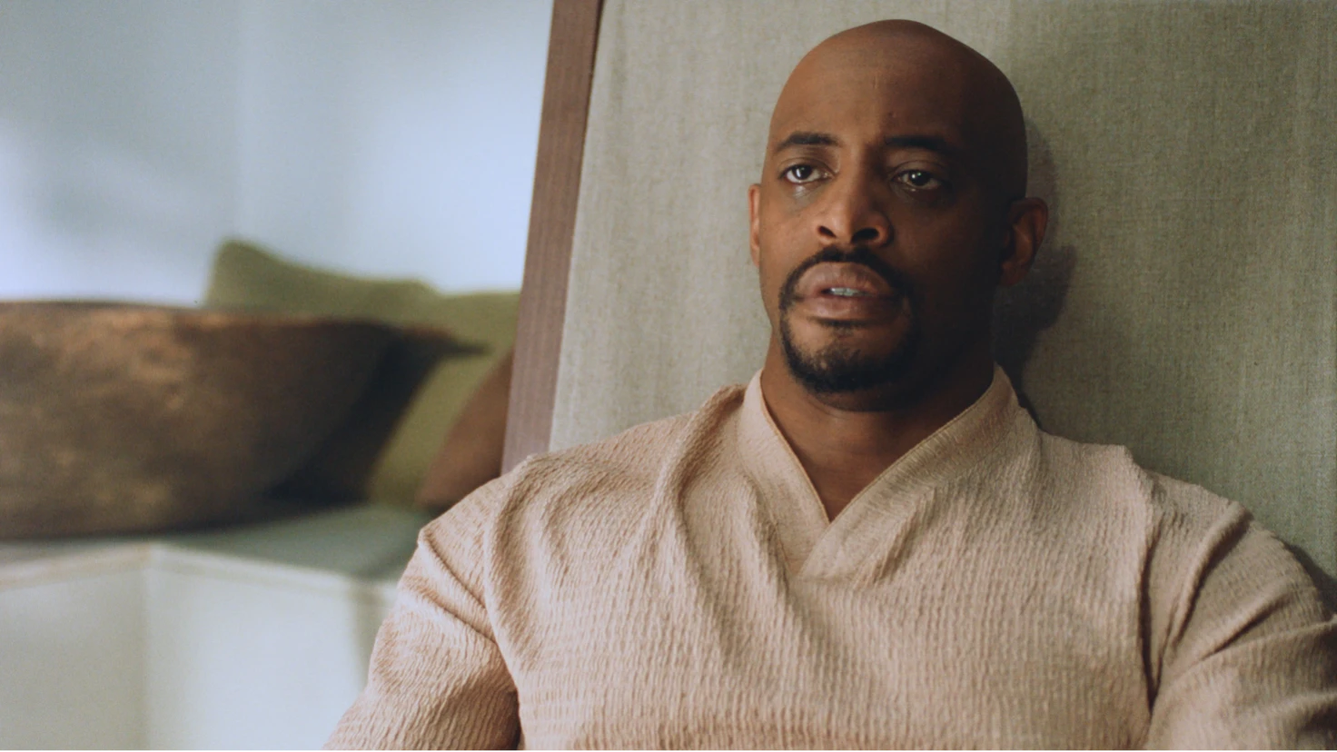 Maleek sits against a soft furnishing, wearing a cream knit top. He has a shaved head and beard, and his expression is contemplative and emotional. Natural light falls across his face.