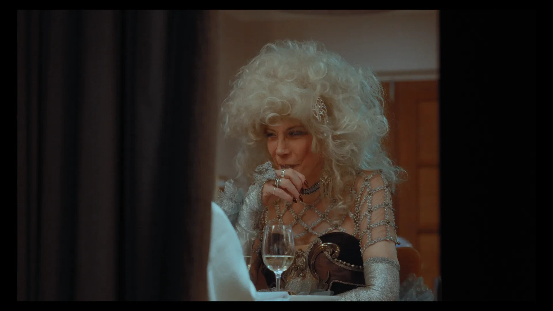 Martine Rose, in a white curly wig and beaded dress, sits at a dimly lit table with a glass of wine.