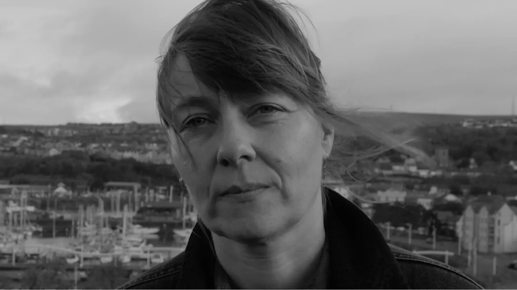 A black and white image of a woman looking at you with a serious face and windswept short hair, with Whitehaven harbour in the background