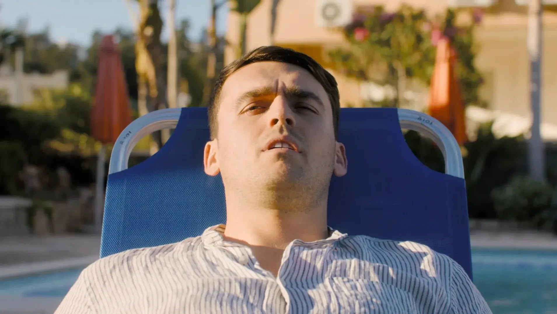Man wearing shirt exhausted on a blue coloured lounger, in a warm country. Sun umbrellas, pool and palm trees out of focus in the background. The sunlight hits him from the left-hand side.