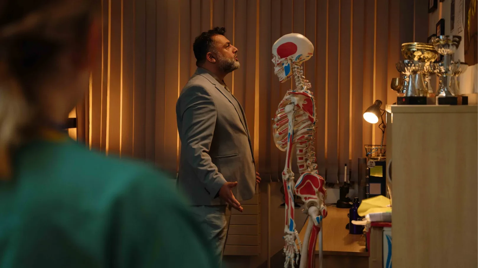 A man (Irvine Iqbal) sings at a medical skeleton watched by another doctor (Annabella Jennings) 